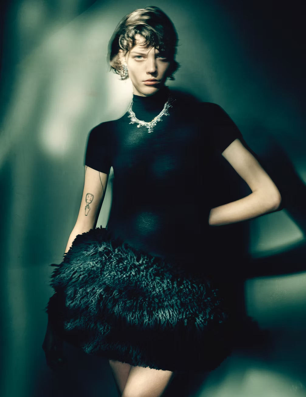 A woman with short hair stands with her hand on her hip, wearing a black dress and jewelry in a studio setting with soft lighting.