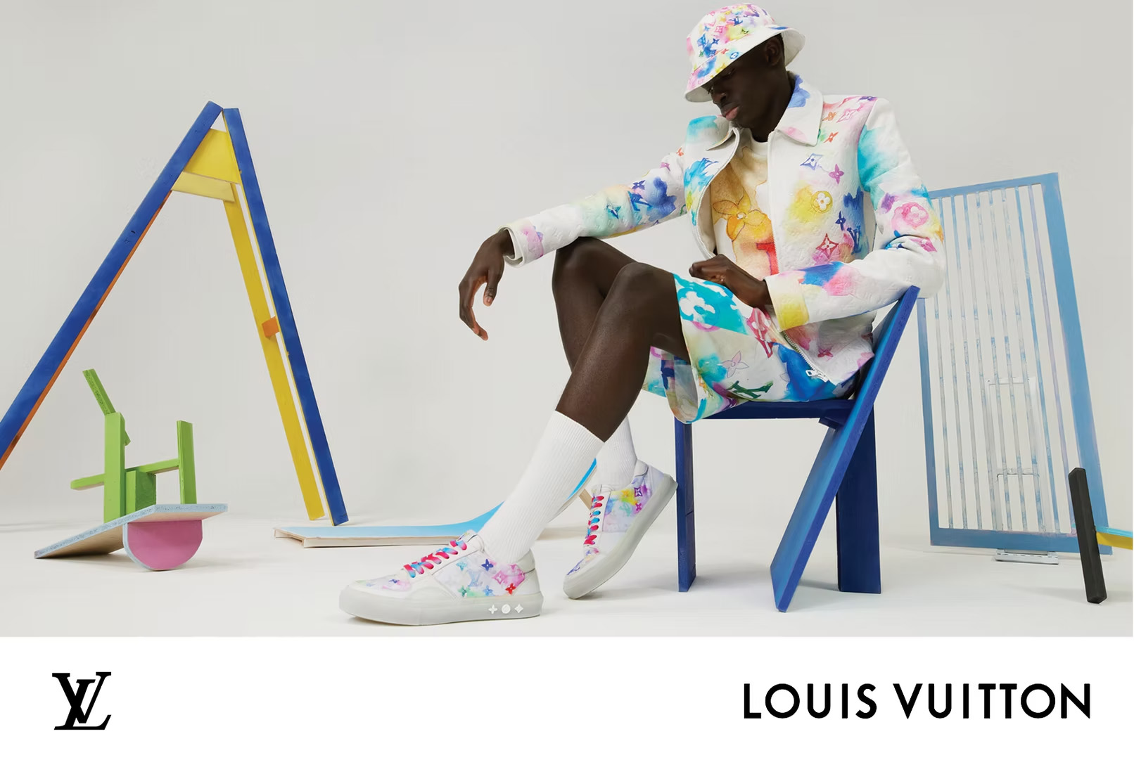 Fashion shot of a Black male model sitting on a blue chair in a colorful jacket, shorts, hat and sneakers in a white studio.