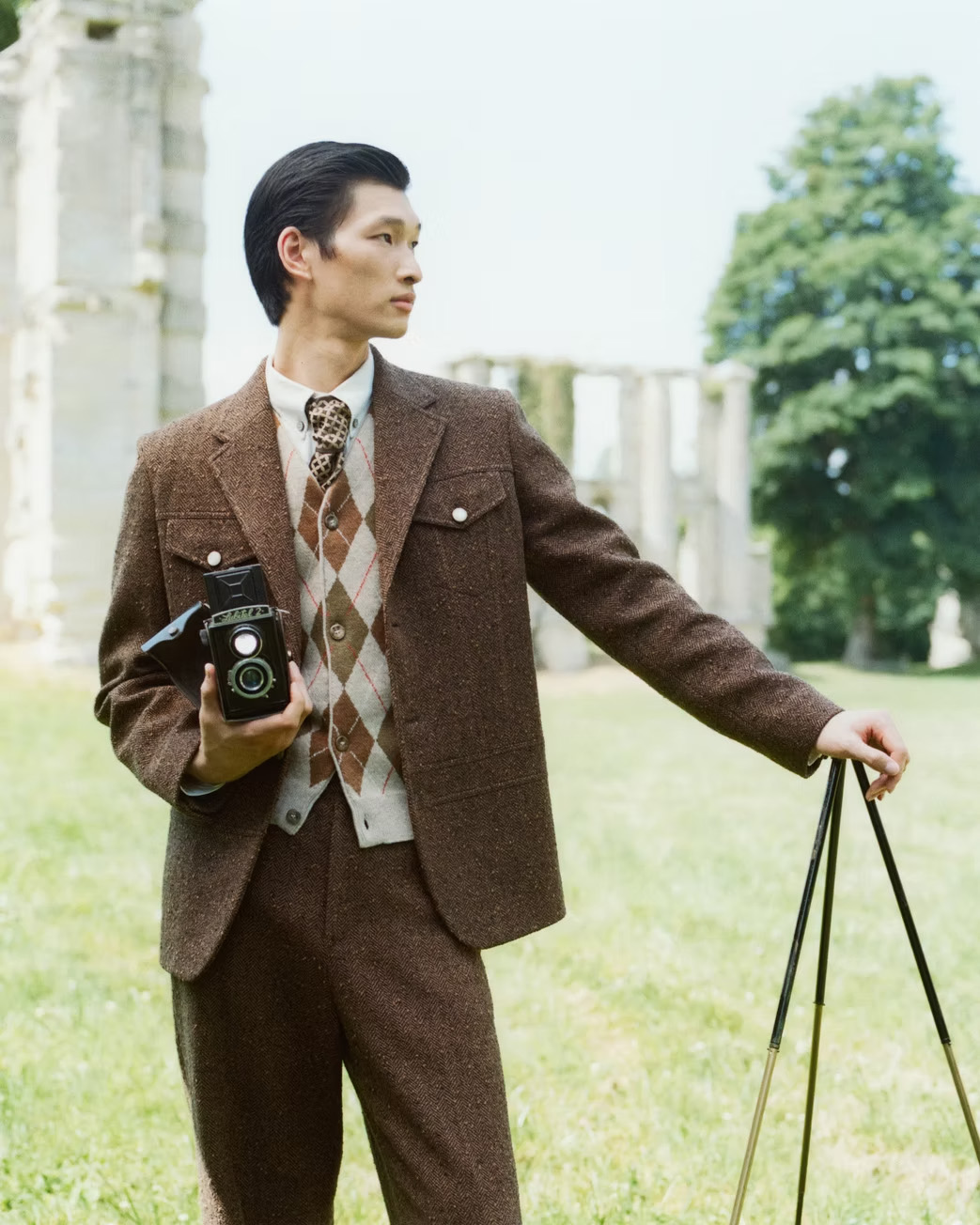 Man in brown suit and argyle vest holds a vintage camera outdoors in a field with distant stone ruins and trees in the background.