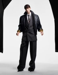 Model in black leather bomber jacket, grey suit jacket, trousers, white shirt, and tie against white background