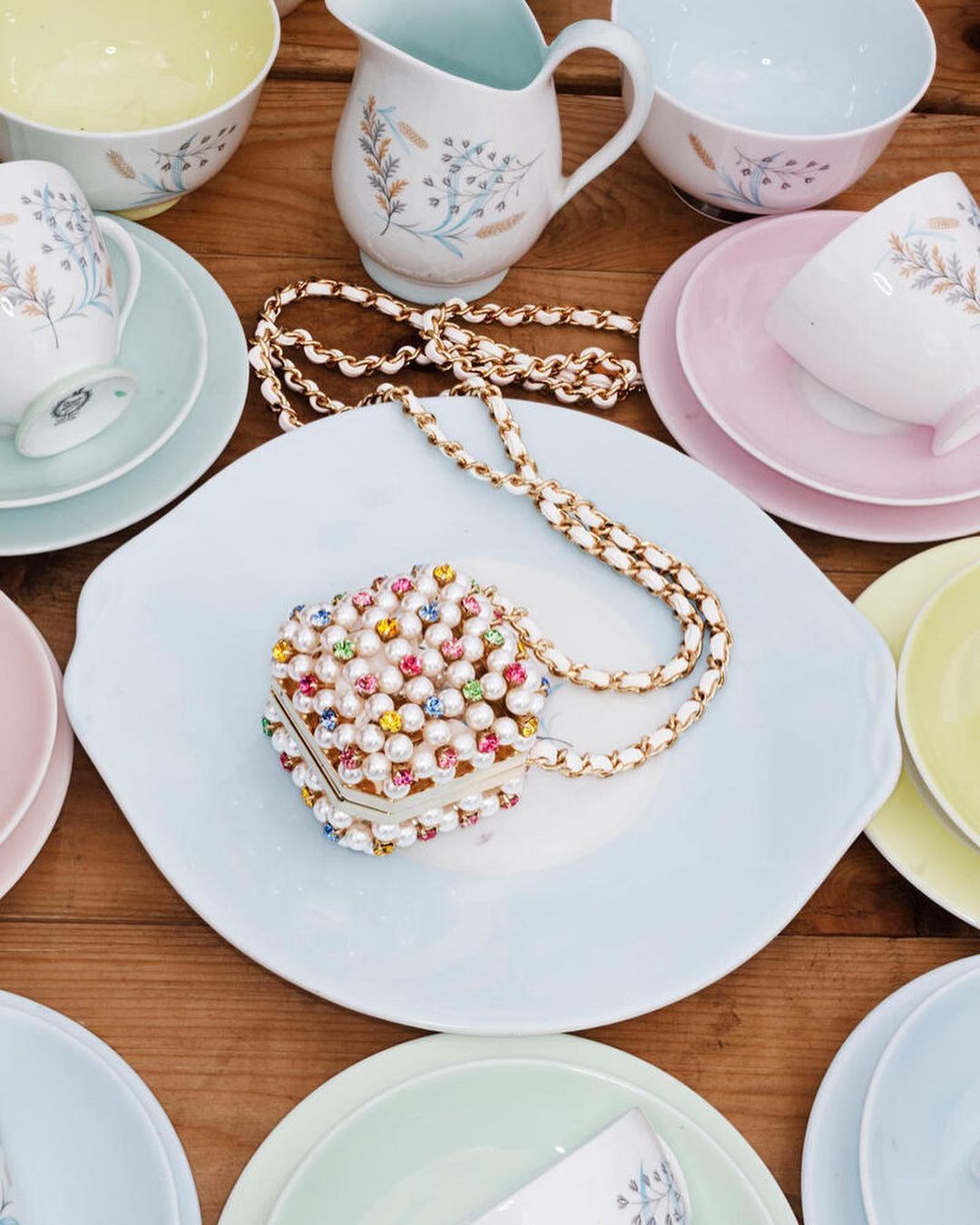 Overhead view of a pearl-beaded purse on a light blue plate, surrounded by colorful vintage floral teacups and serving pieces on wood.