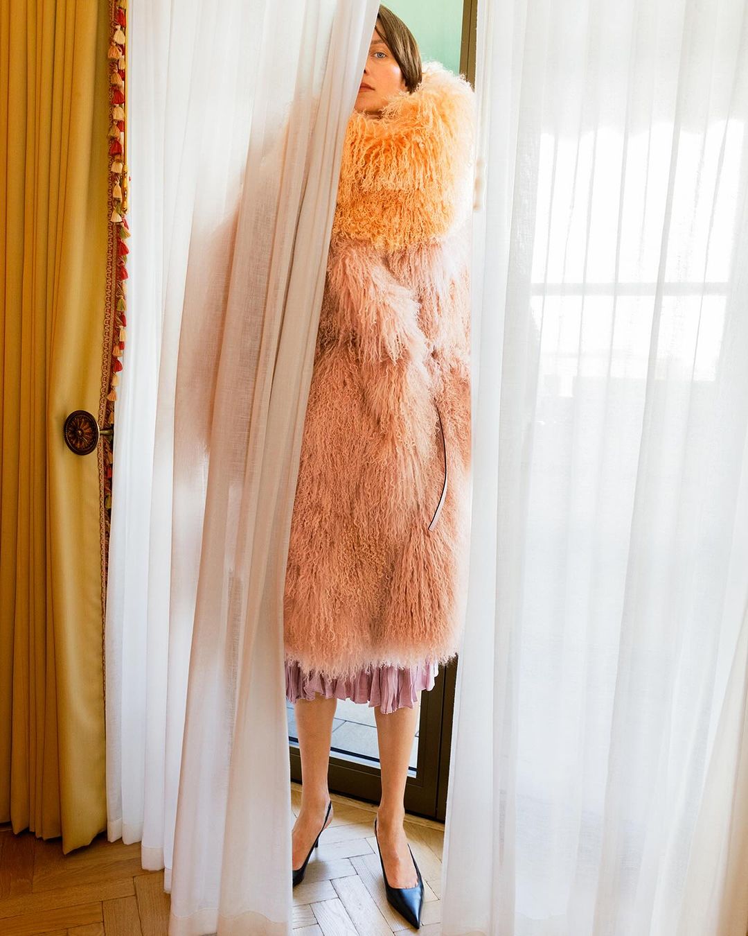 Adult female stands behind sheer curtains wearing orange and pink fur coat, pleated skirt and black high heels indoors near window.