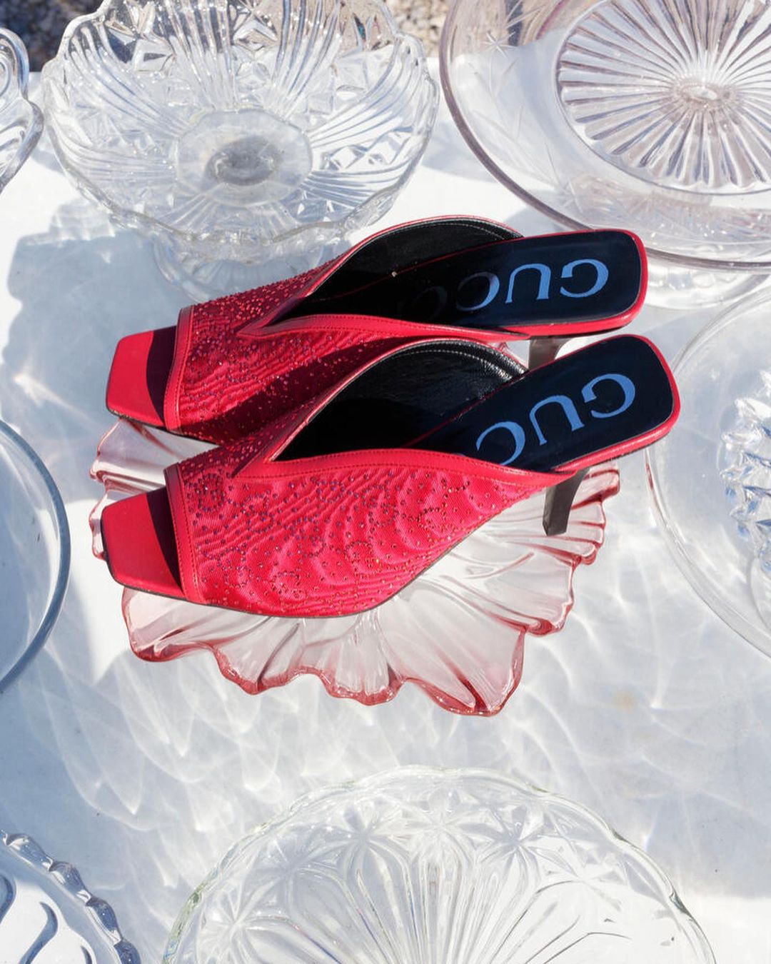 Elevated view of a pair of red Gucci heeled sandals on a light pink glass dish, surrounded by assorted clear glass dishes and bowls.
