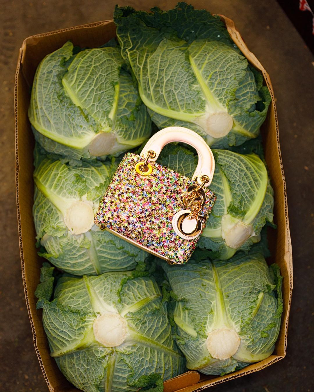 Box filled with green cabbages and a colorful sequined handbag with a white handle and gold chain. Still life, overhead shot.