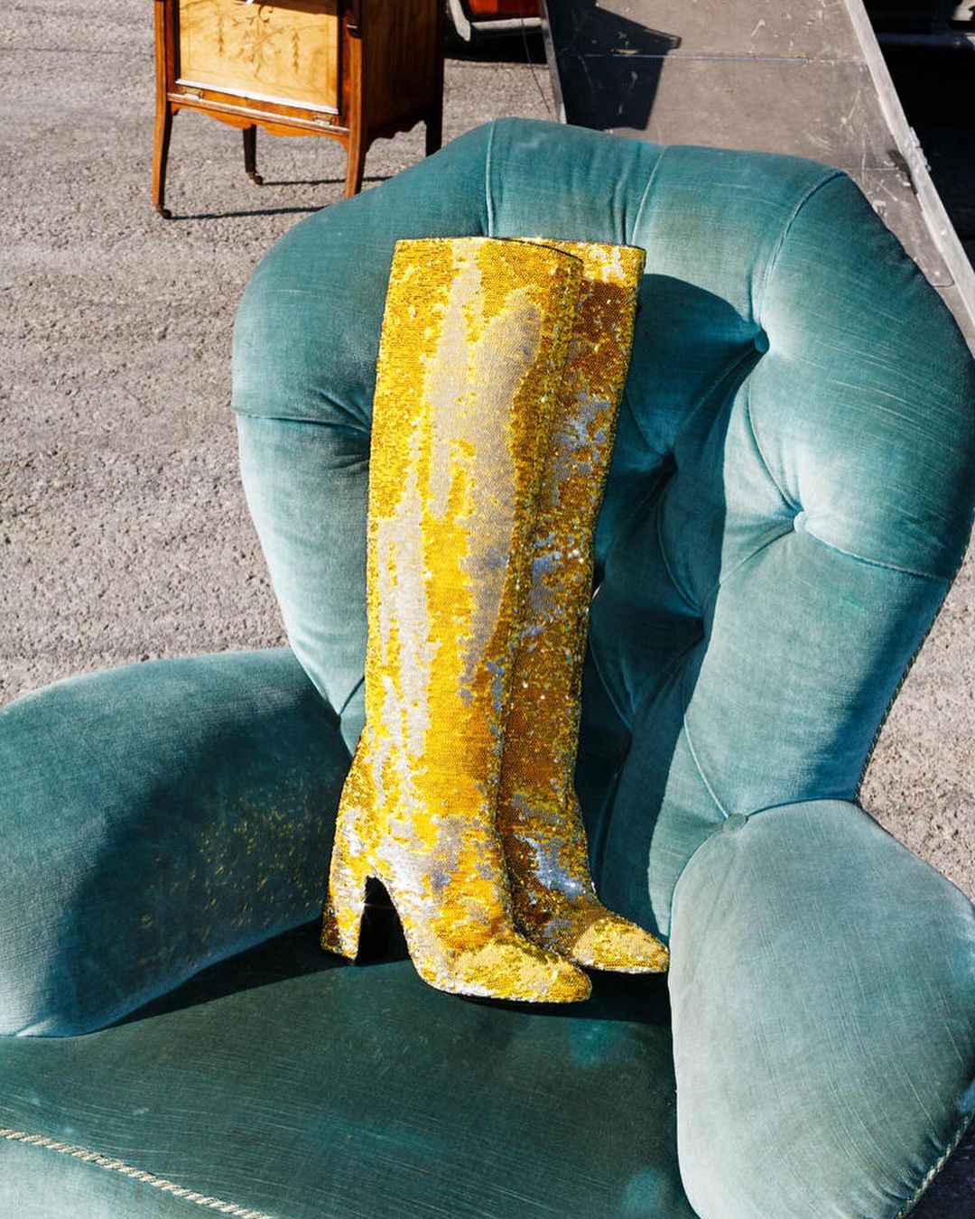 Sequin-covered gold high-heel boots rest on a green velvet armchair, with a vintage wooden cabinet and concrete visible in the background.