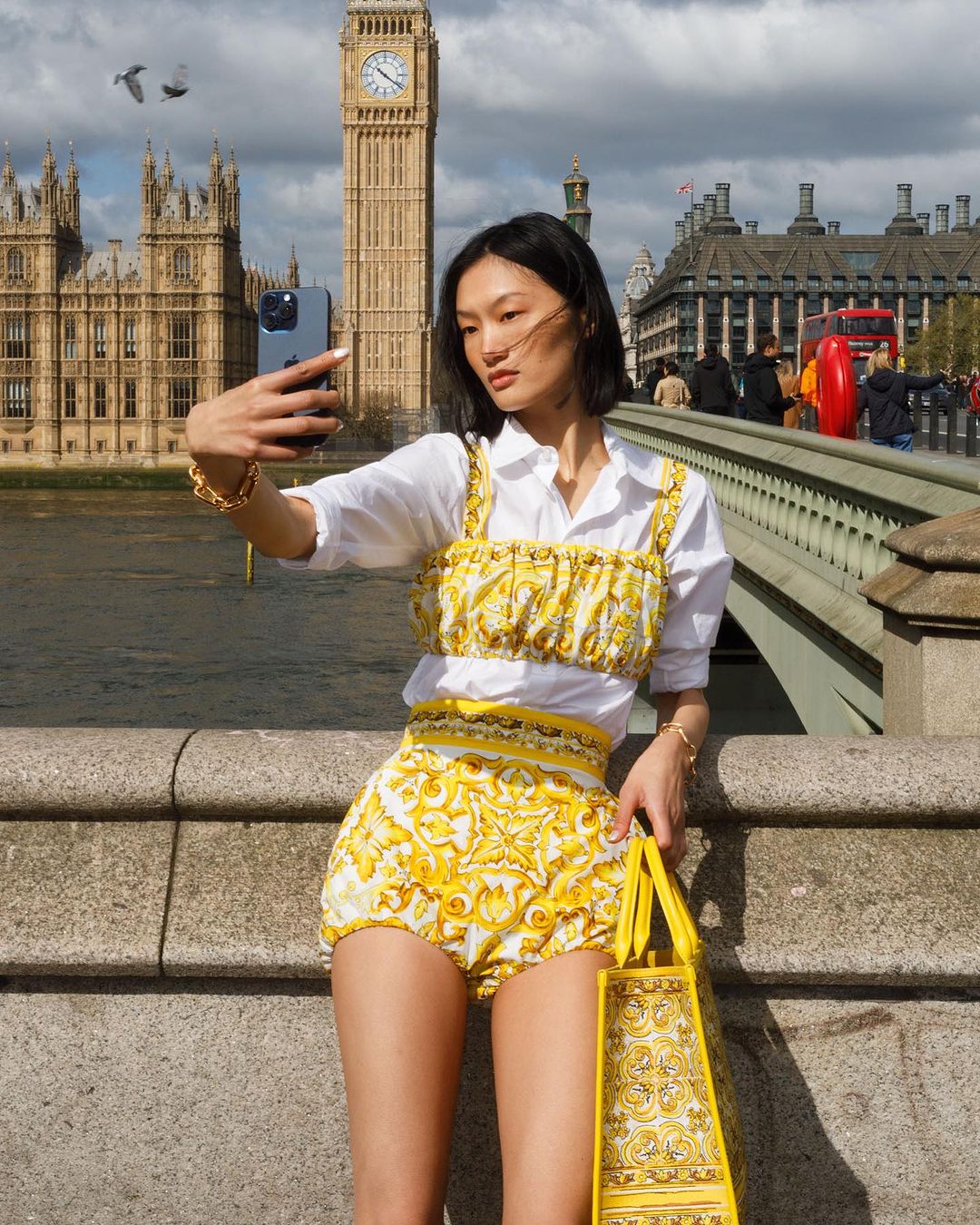 Asian woman takes selfie in London with Big Ben in the background. She wears a yellow patterned outfit with a white shirt and carries a tote.