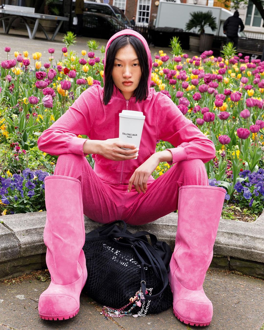 Woman in pink tracksuit & boots sits amid tulips, holding coffee cup by Balenciaga, outside with building and car in the background.