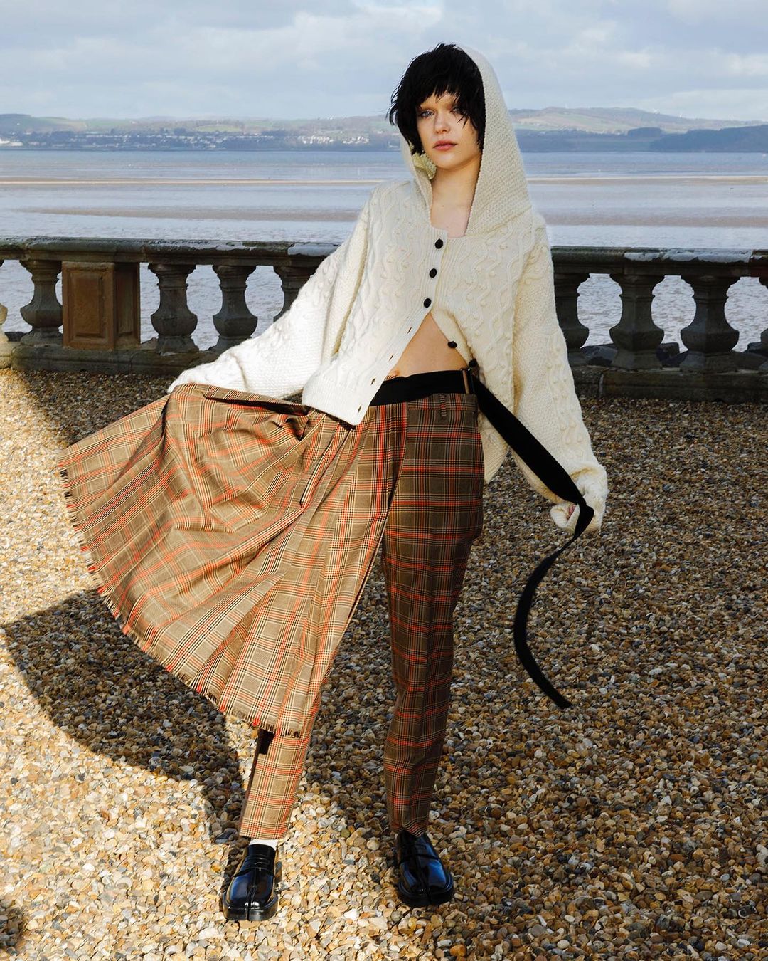 Fashionable young adult female poses outdoors in a cream cardigan, plaid skirt and pants, and patent leather shoes near the water.