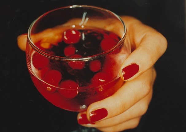 Close-up of a woman's hand holding a clear glass filled with a red liquid and multiple red cherries against a dark background.