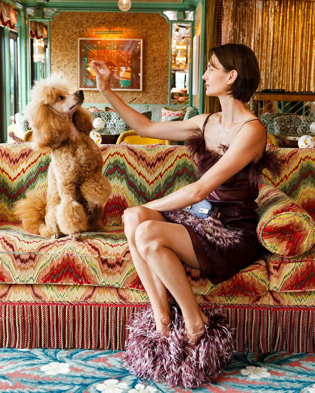 Woman sits on patterned couch with her tan poodle. She's wearing a feathered mini dress and feathered high heels in an indoor setting.