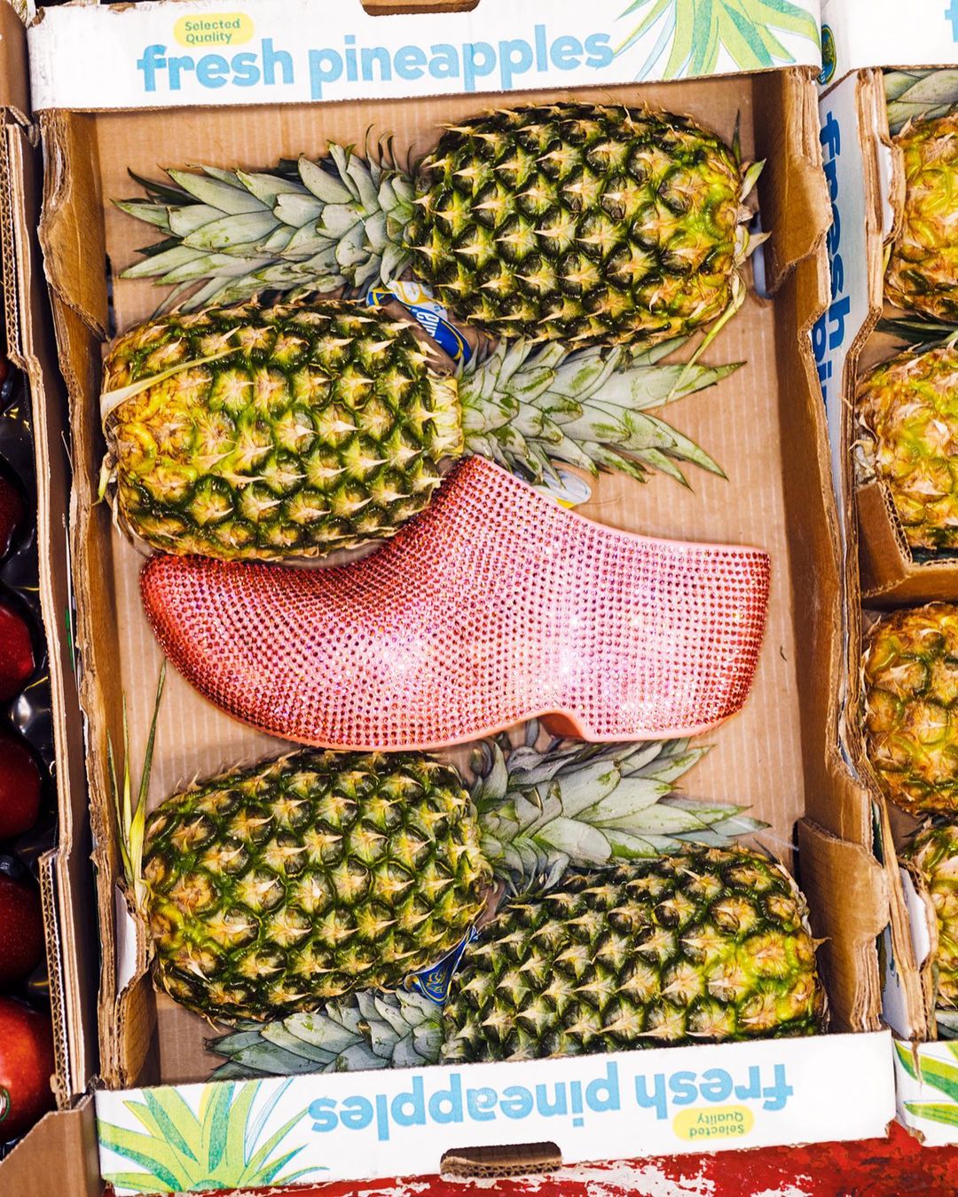Close-up of pineapples in a cardboard box, with a bright pink and red traditional Dutch wooden shoe nestled among the fruit.