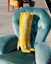 A pair of gold glitter knee-high boots resting upright in a tufted turquoise velvet armchair outdoors.