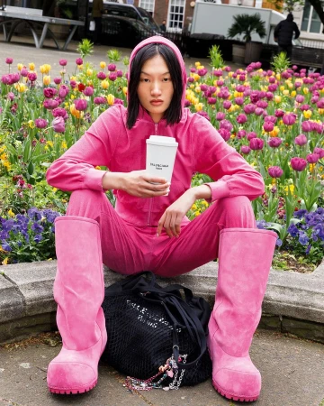 Asian woman in pink tracksuit and boots holding a white cup, seated in front of a colorful flower bed in London by Martin Parr.