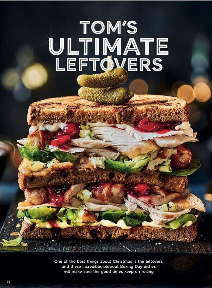 Stacked sandwich with turkey, sausage, greens, cranberries, pickles on dark wood surface, illuminated by overhead lighting, against dark bokeh background.