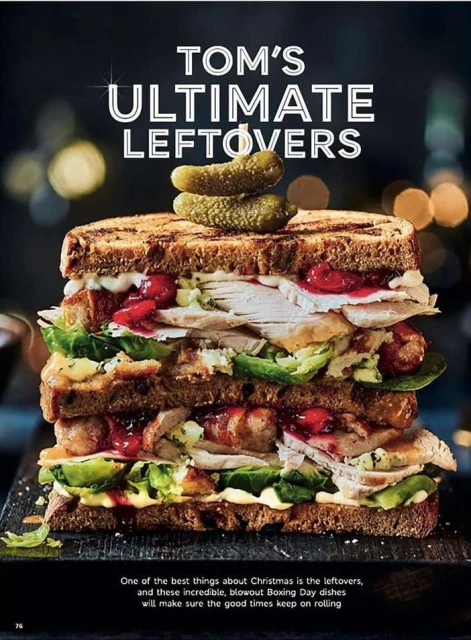 Stacked sandwich with turkey, sausage, greens, cranberries, pickles on dark wood surface, illuminated by overhead lighting, against dark bokeh background.