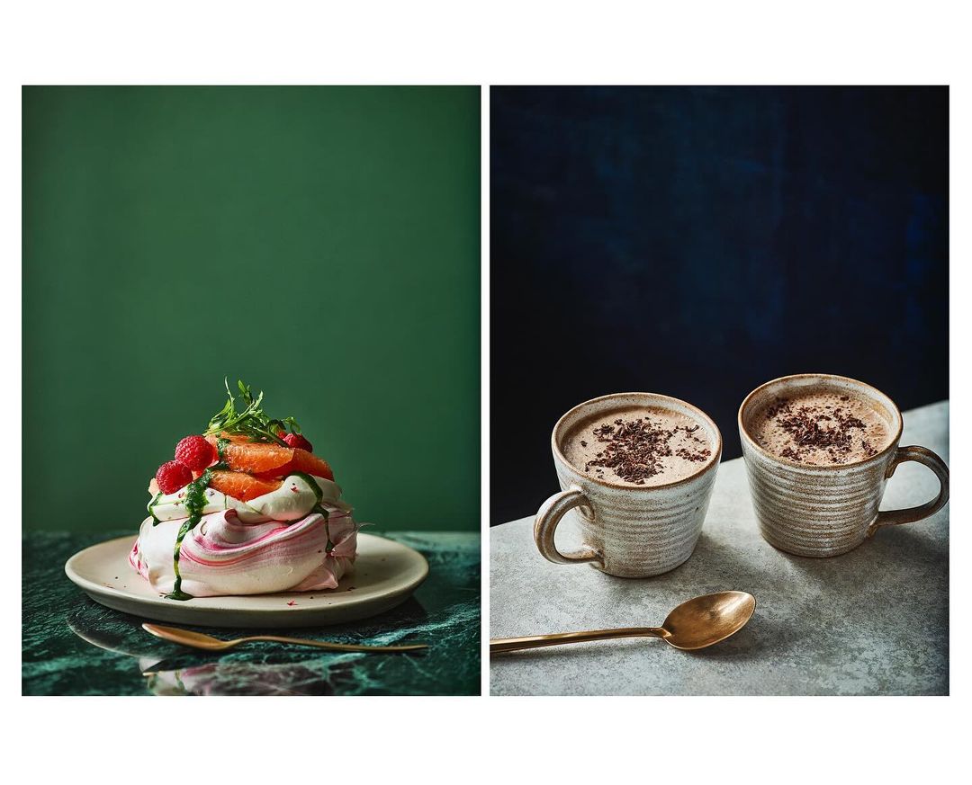 Eye-level view displays two separate scenes: a pavlova dessert with fruit and two mugs of cocoa or coffee with a brass spoon.