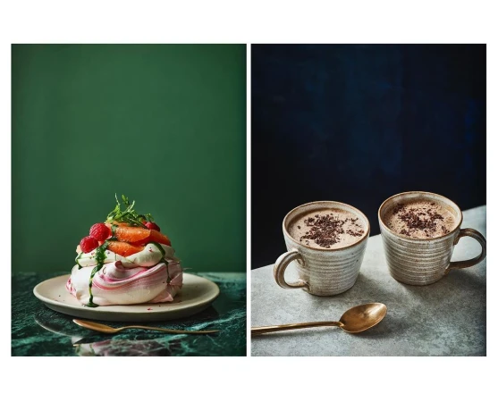 Eye-level view displays two separate scenes: a pavlova dessert with fruit and two mugs of cocoa or coffee with a brass spoon.