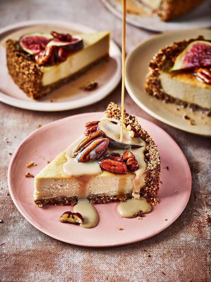 Close-up of three slices of cheesecake topped with figs and nuts with caramel sauce being poured, set on pink and off-white plates.