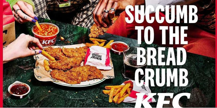 Overhead shot of fried chicken, fries, and beans on a table, alongside hands reaching for the food; KFC branding is visible.