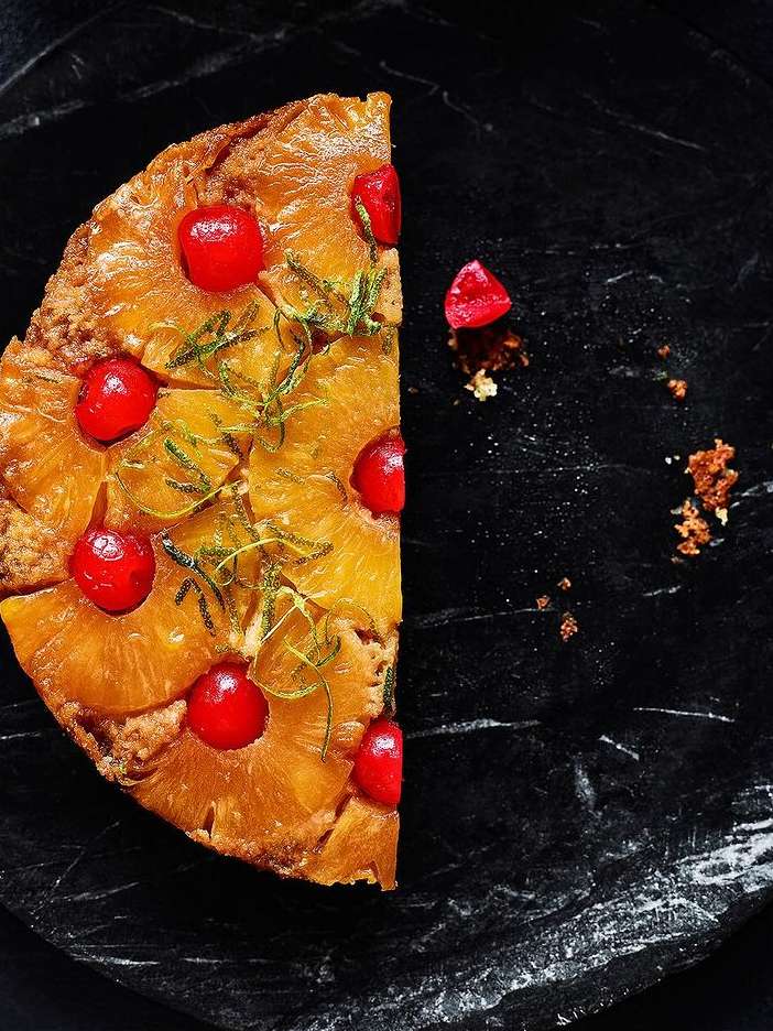 Overhead shot of a slice of pineapple upside-down cake with cherries, arranged on a black marble surface with crumbs nearby.