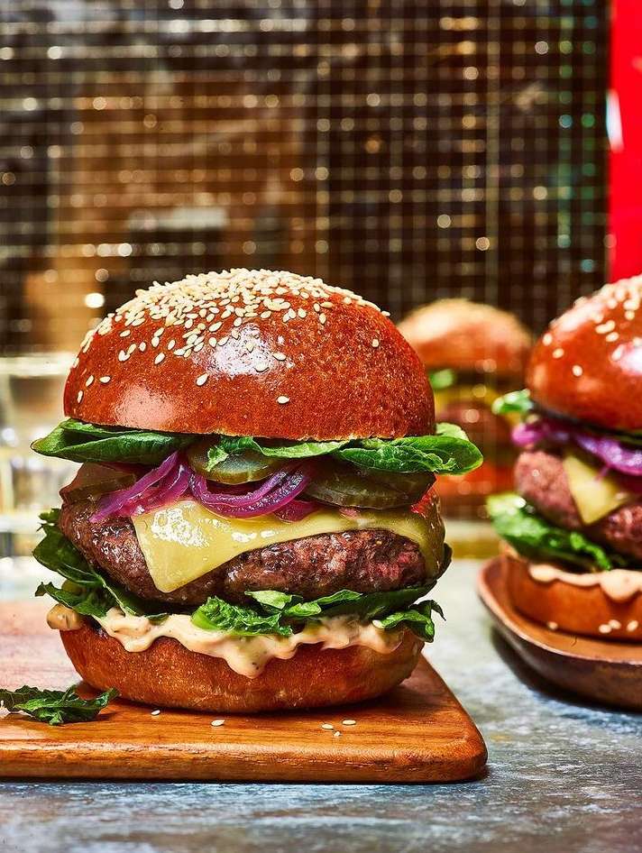 Three gourmet burgers with beef patties, melted cheese, pickles, lettuce, and red onion on sesame buns served on wood boards.