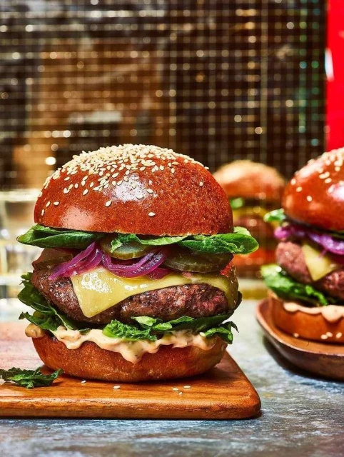 Three gourmet burgers with beef patties, melted cheese, pickles, lettuce, and red onion on sesame buns served on wood boards.
