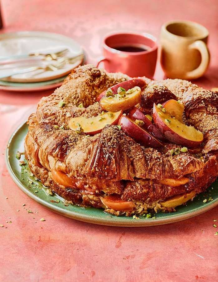 Overhead shot of a croissant cake with peaches and pistachios on a green plate, accompanied by coffee cups, creating an inviting food still life.