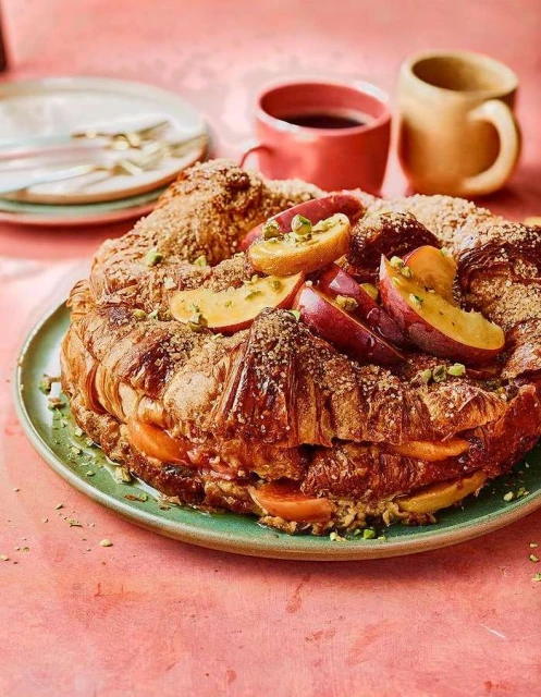 Overhead shot of a croissant cake with peaches and pistachios on a green plate, accompanied by coffee cups, creating an inviting food still life.
