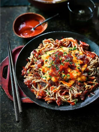 Overhead shot of noodles with bacon and orange sauce in a black bowl, accompanied by chopsticks and a bowl of red sauce.