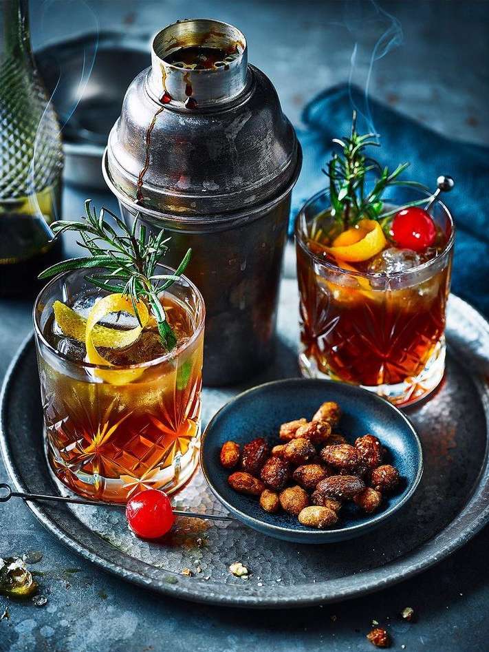 Two cocktails with garnishes, a cocktail shaker, and a bowl of nuts are arranged on a metallic tray in a moody, inviting atmosphere.