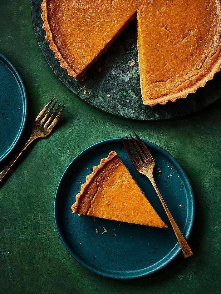 Overhead shot of a sliced pumpkin tart on a blue plate with gold forks, with the rest of the tart on a green marble surface.