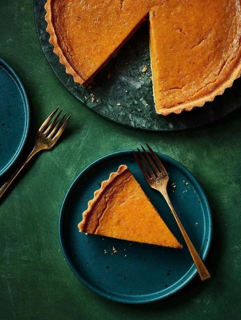 Overhead shot of a sliced pumpkin tart on a blue plate with gold forks, with the rest of the tart on a green marble surface.