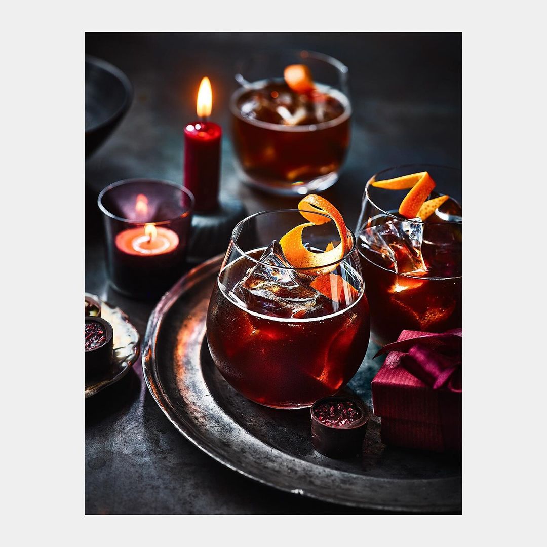 Three cocktail glasses filled with dark liquid and ice, garnished with orange peel, alongside lit candles and small chocolate desserts.