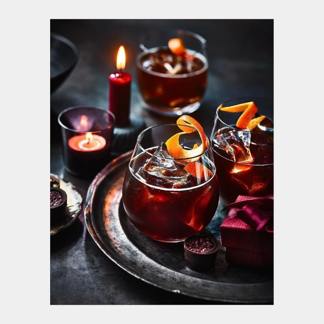 Three cocktail glasses filled with dark liquid and ice, garnished with orange peel, alongside lit candles and small chocolate desserts.