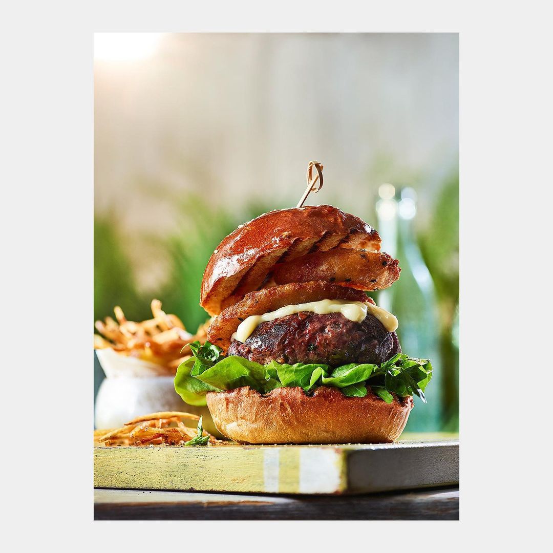 A gourmet burger with onion rings is centered on a wooden board next to a dish of fries, set against a softly blurred, bright backdrop.