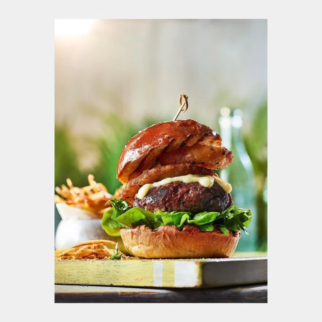A gourmet burger with onion rings is centered on a wooden board next to a dish of fries, set against a softly blurred, bright backdrop.