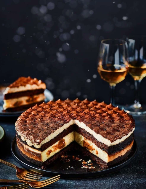 Tiramisu cake on a black plate with golden forks and wine glasses in background, suggesting dessert or celebration. Food photography.