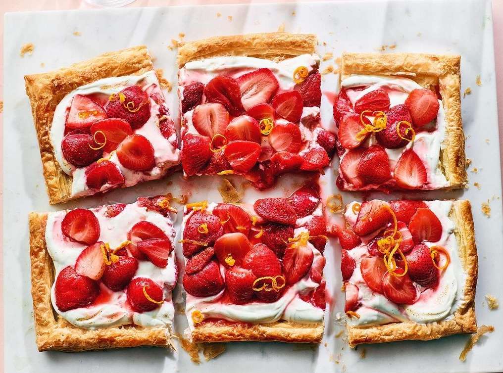 Overhead shot of six square strawberry tarts topped with whipped cream and orange zest on a white marble surface, food photography.