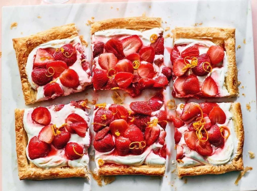 Overhead shot of six square strawberry tarts topped with whipped cream and orange zest on a white marble surface, food photography.