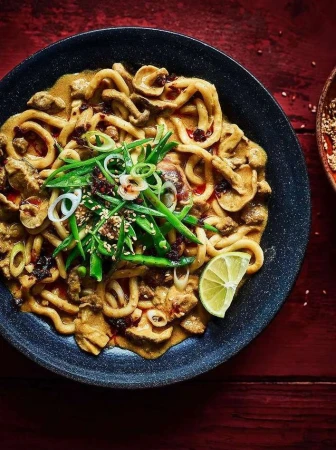 Overhead shot of a bowl filled with noodles and vegetables, garnished with sesame seeds, lime, and green onions, on a wooden surface.