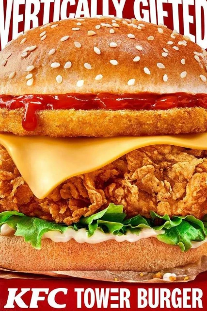 A close-up, eye-level shot of a KFC Tower Burger. The focus is on the layers of fried chicken, cheese, lettuce, and sauce, creating an appetizing image.