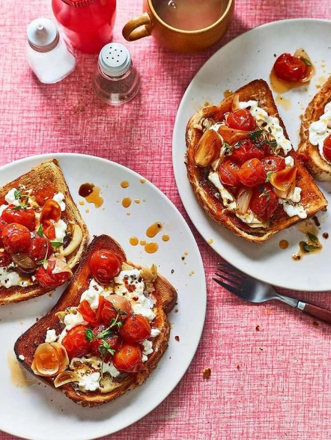Overhead shot of two plates of toasted bread topped with tomatoes, garlic, and cheese, arranged on a pink tablecloth, with salt shakers.