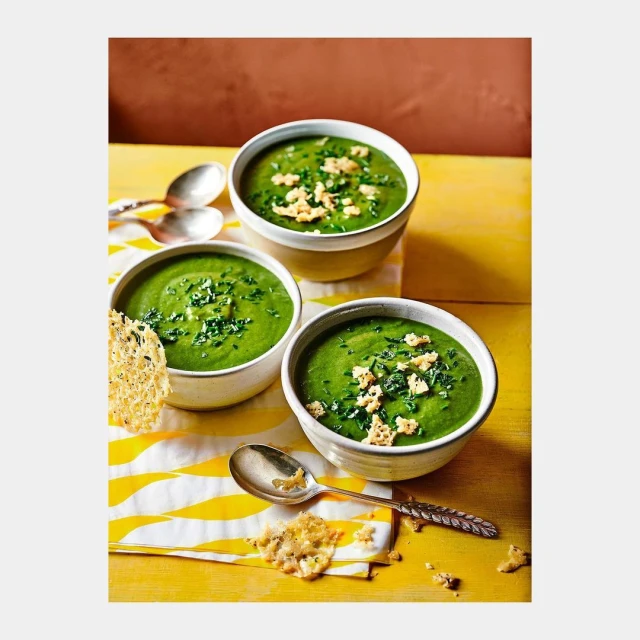 Three bowls of vibrant green soup topped with herbs and croutons sit on a yellow striped cloth next to silver spoons, food photography.