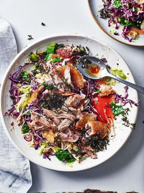 Overhead shot of a white bowl filled with shredded meat, red cabbage, bean sprouts, greens, and a silver spoon with remnants of sauce.