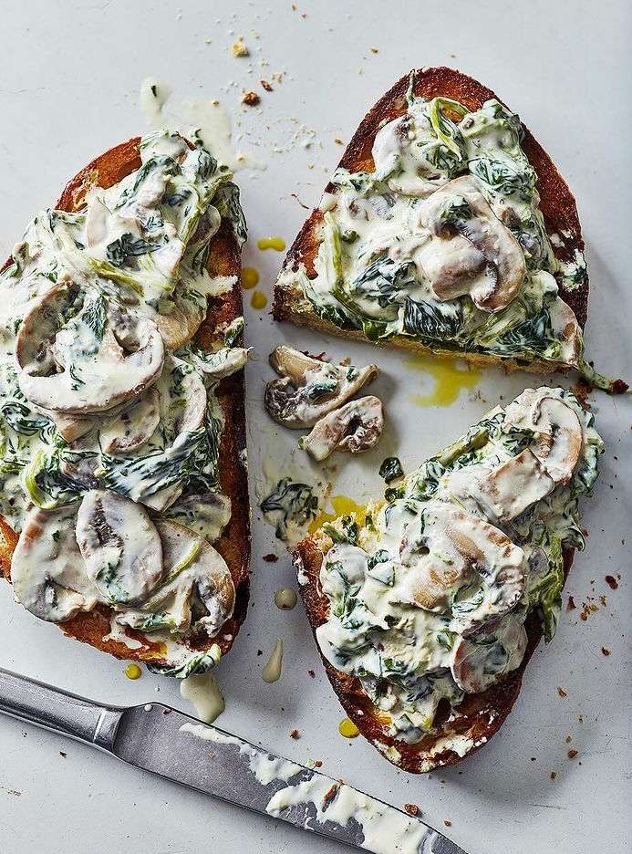 Two slices of toasted bread topped with creamy spinach, sliced mushrooms, and a drizzle of olive oil, served with a knife.