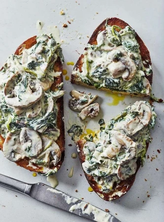 Two slices of toasted bread topped with creamy spinach, sliced mushrooms, and a drizzle of olive oil, served with a knife.