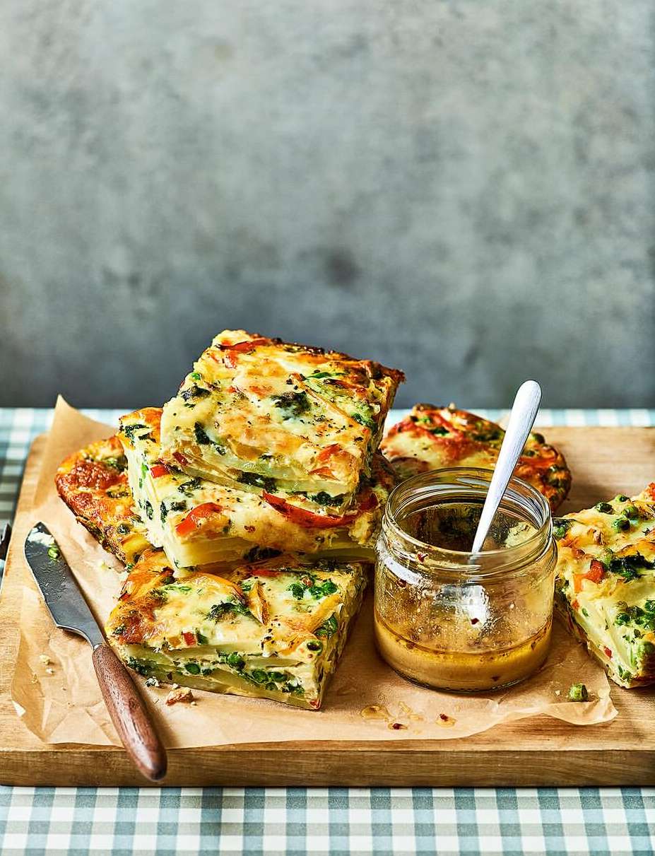 Overhead shot of savory vegetable frittata squares stacked on a wooden board next to a jar of sauce and a knife.