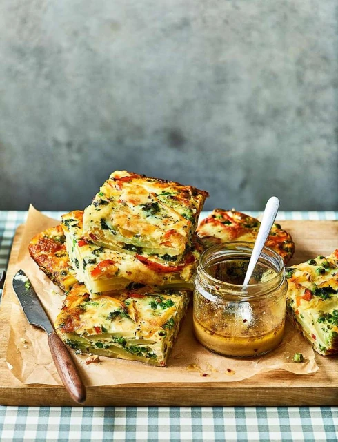 Overhead shot of savory vegetable frittata squares stacked on a wooden board next to a jar of sauce and a knife.