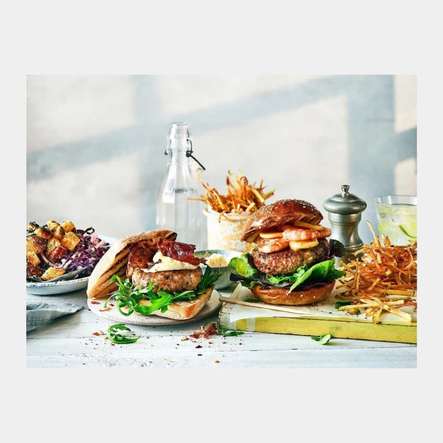 Two gourmet burgers topped with shrimp and bacon sit on a white table, alongside a water bottle, assorted side dishes, and garnishes.