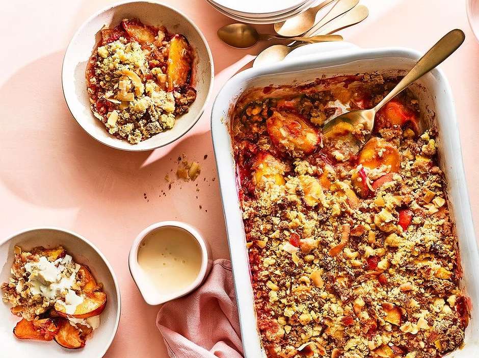 Overhead shot of a peach cobbler baking dish with two bowls, golden spoons, a white creamer, pink linen, and a peach background.